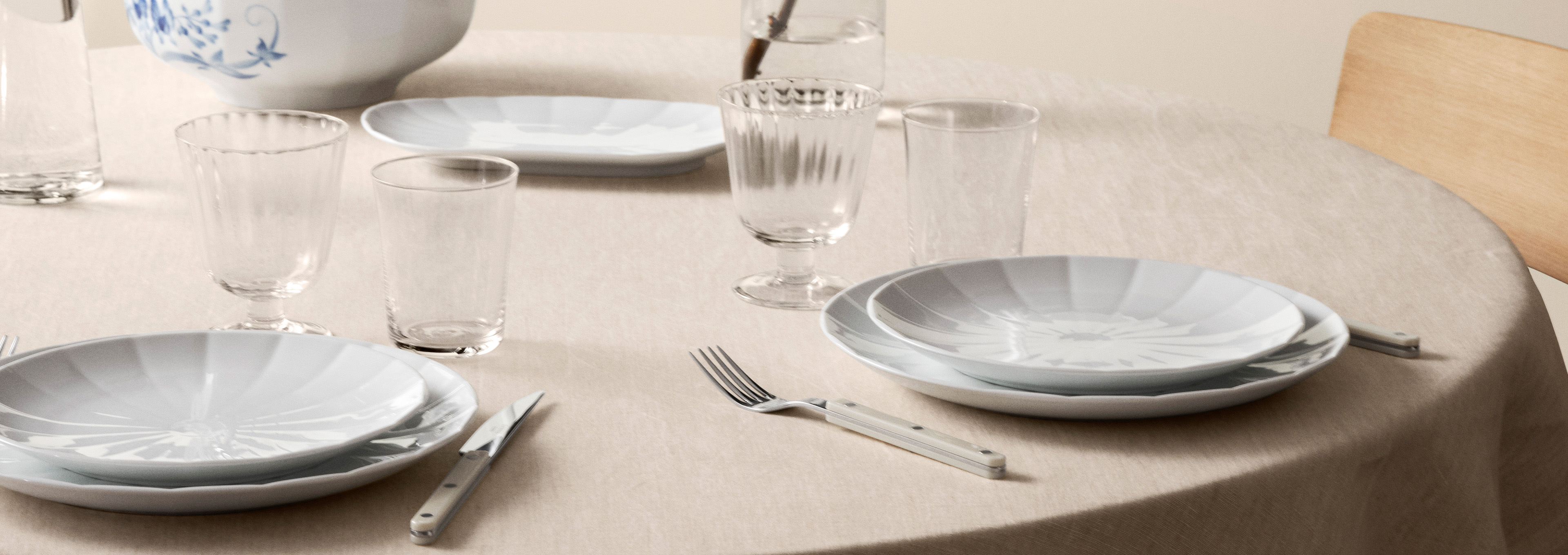 Et bord med en beige dug dækket til to med Hvid Facet middagstallerkener